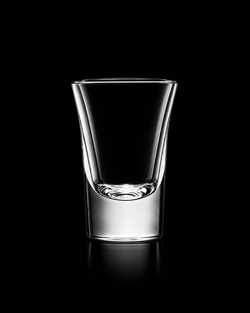 A clear glass shot glass with a wide rim and a thick base, photographed on a solid black background with a soft reflection below.