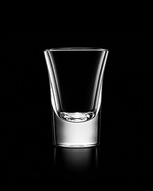A clear glass shot glass with a wide rim and a thick base, photographed on a solid black background with a soft reflection below.