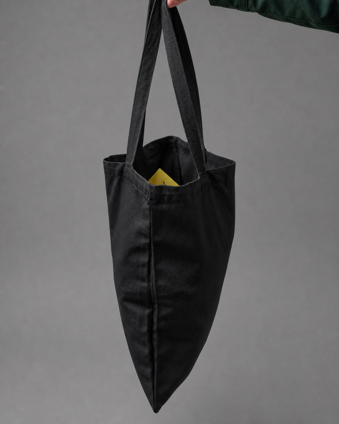 A black canvas tote bag with long shoulder straps, held by a hand against a solid gray studio background.