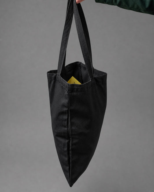 A black canvas tote bag with long shoulder straps, held by a hand against a solid gray studio background.